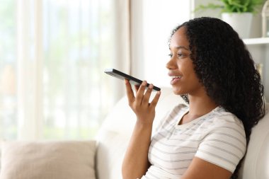 Happy black woman dictating message on phone sitting on a sofa at home clipart