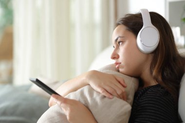 Üzgün kadın telefon ve kulaklıkla müzik dinliyor. Evdeki pencereden bakıyor.