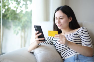 Ciddi Asyalı kadın kredi kartı ve akıllı telefonuyla internetten alışveriş yapıyor.