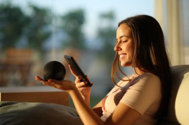 Gün batımında akıllı hoparlörü cep telefonuyla eşleştiren mutlu kadın.