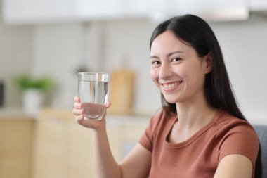 Mutfakta elinde bir bardak su tutan mutlu Asyalı kadın kameraya bakıyor.