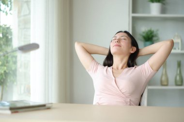 Asyalı bir kadın evinde bir odada, kolları kafasında bir sandalyede oturup rahatlıyor.