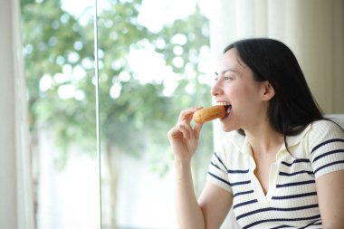 Mutlu Asyalı kadın donut ısırıyor. Evinin penceresinden içeri bakıyor.