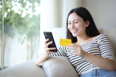 Mutlu Asyalı kadın evde kredi kartı ve cep telefonuyla online alışveriş yapıyor.