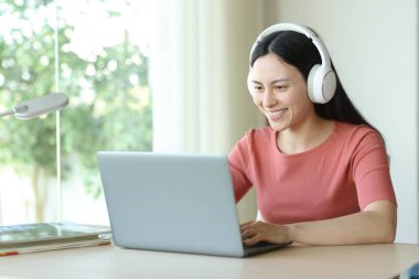 Evdeki bir masada dizüstü bilgisayar kullanan kulaklıklı mutlu Asyalı kadın.