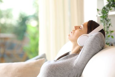 Evde bir kanepede oturmuş müzik dinleyen rahat yetişkin bir kadının yan görüntüsü.