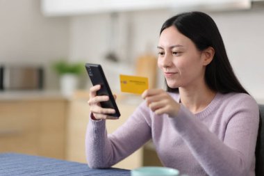 Ciddi Asyalı bir kadın evdeki mutfakta kredi kartı ve cep telefonuyla online alışveriş yapıyor.