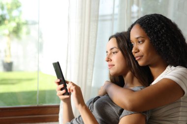 Ciddi ırklar arası lezbiyen çift evdeki pencerenin yanında akıllı telefonları kontrol ediyor.