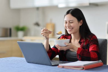 Mutlu Asyalı öğrenci laptopuyla evde kahvaltıda mısır gevreği yiyor.