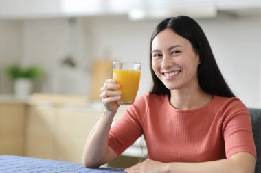 Mutlu Asyalı kadın portakal suyu bardağı gösteriyor. Evdeki mutfakta kameraya bakıyor.