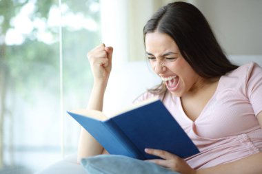 Evdeki koltukta kitap okuyan heyecanlı bir kadın.