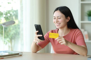 Mutlu Asyalı kadın internetten kredi kartı ve telefonuyla evdeki bir masada alışveriş yapıyor.