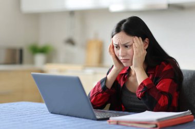 Endişeli Asyalı öğrenci bilgisayarın içeriğini kontrol ediyor ve evdeki mutfakta şikayet ediyor.