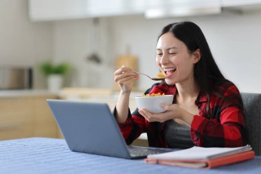 Mutlu Asyalı öğrenci laptopuyla evde kahvaltıda mısır gevreği yiyor.