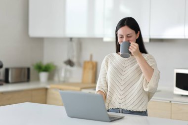 Asyalı kadın kahve içiyor ve dizüstü bilgisayarını kontrol ediyor. Mutfakta duruyor.