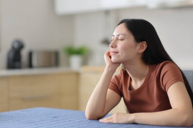 Asyalı kadın evde mutfakta oturmuş gözleri kapalı dinleniyor.