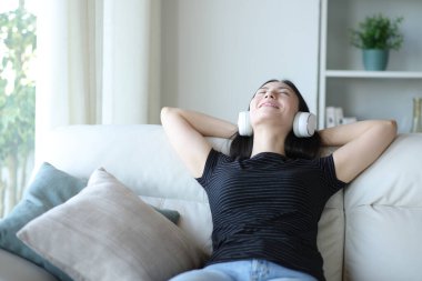 Mutlu Asyalı kadın evde bir kanepede oturmuş kulaklıkla müzik dinliyor.