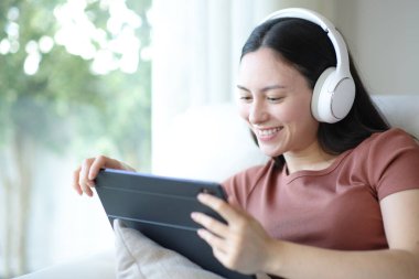 Kulaklıklı mutlu Asyalı kadın tablette video seyrediyor evdeki kanepede oturuyor.