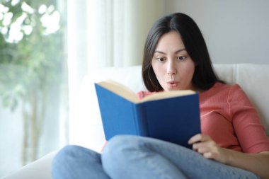 Evdeki kanepede oturan Asyalı kadın kitap okuyor.