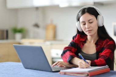 Bilgisayarı ve kulaklığı olan Asyalı bir öğrenci evde mutfakta notlar alıyor.