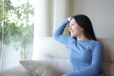 Mutlu Asyalı kadın evdeki kanepede oturup pencereden bakıyor.
