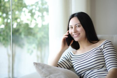 Telefonda konuşan mutlu Asyalı kadın evdeki koltukta oturan kameraya bakıyor.