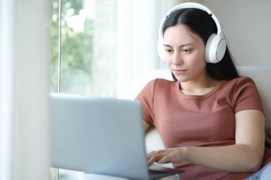 Kulaklık takan Asyalı bir kadın evdeki koltukta oturuyor.