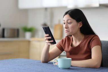 Kafası karışmış Asyalı kadın mutfaktaki telefon içeriğini kontrol ediyor.