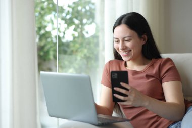 Mutlu Asyalı kadın evdeki koltukta birden fazla cihaz kullanıyor.