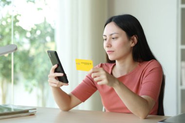 Ciddi Asyalı kadın evde bir odada kredi kartı ve telefon kullanarak internetten satın alıyor.