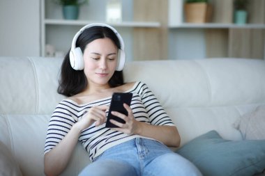 Asyalı kadın telefondaki medya içeriğini izliyor evdeki kanepede oturuyor.