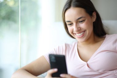 Mutlu kadın cep telefonu kullanıyor. Gülümsüyor. Evdeki koltukta oturuyor.