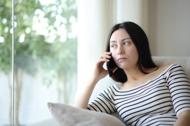 Sıkıcı Asyalı kadın telefonda konuşurken evde bir kanepede oturuyor.