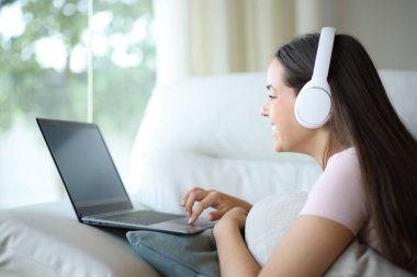 Dizüstü bilgisayar kullanan mutlu bir kadın. Kulaklık takıyor. Evdeki bir kanepede yatıyor.