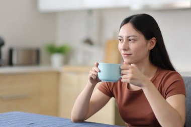 Rahatlamış Asyalı kadın evdeki mutfakta içiyor ve düşünüyor.