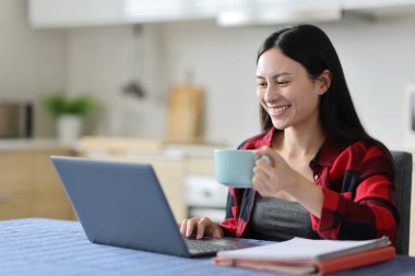 Mutlu Asyalı kadın evdeki mutfakta dizüstü bilgisayar kullanarak içiyor ve e-öğreniyor.