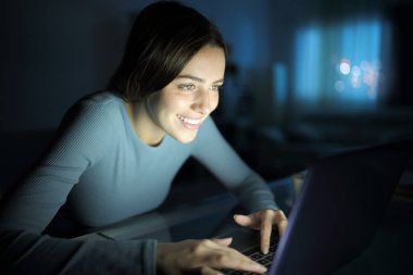Gece evde masa başında dizüstü bilgisayar kullanan mutlu bir kadın.