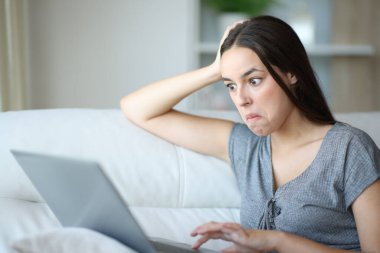 Kafası karışmış bir kadın evdeki bir kanepede oturmuş dizüstü bilgisayarını kontrol ediyor.