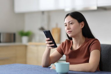 Dikkati dağılmış Asyalı kadın telefonu elinde tutuyor ve evdeki mutfağa bakıyor.