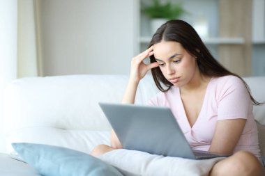 Evdeki bir koltukta dizüstü bilgisayar kullanan şaşkına dönmüş bir kadın.