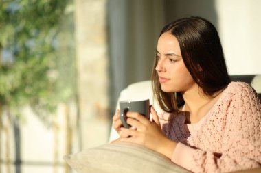 Ciddi ciddi kahve içen düşünceli bir kadın evdeki pencereden dışarı bakıyor.