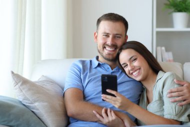 Mutlu bir çift telefonlarını tutuyor ve sana bakıyor. Evdeki koltukta oturuyor.