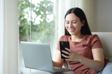 Bir kanepede oturan mutlu Asyalı kadın telefon ve laptop kullanıyor.