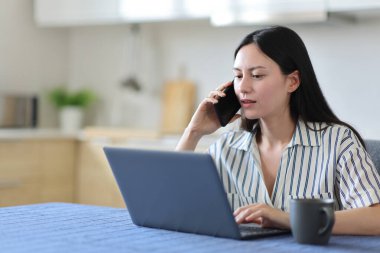 Asyalı kadın dizüstü bilgisayarını kontrol ediyor. Mutfakta telefonla konuşuyor.