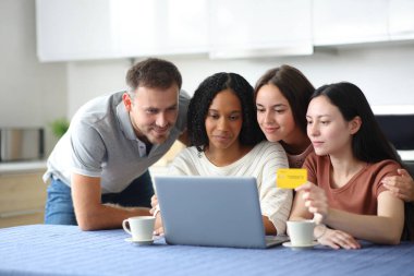 Dört farklı ırktan arkadaş, evdeki mutfaktan kredi kartı ve dizüstü bilgisayarla online alışveriş yapıyor.