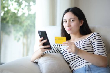 İnternetten alışveriş yapan ciddi Asyalı kadın evdeki kanepede oturmuş kameraya bakıyor.