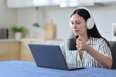 Laptop ve kulaklık kullanan Asyalı bir kadın mutfakta kahve içiyor.