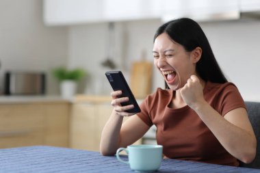 Heyecanlı Asyalı kadın evdeki mutfakta telefon kullanıyor.