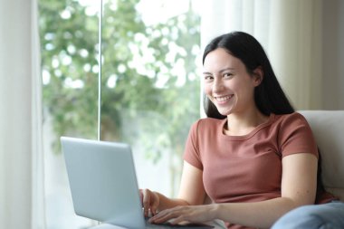 Mutlu Asyalı kadın dizüstü bilgisayar kullanıyor. Kameraya bakıyor. Evdeki koltukta oturuyor.