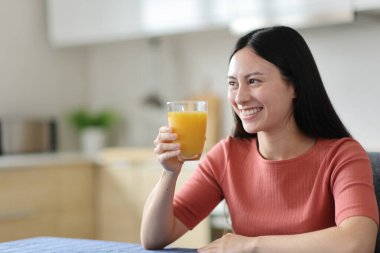 Mutlu Asyalı kadın portakal suyu içiyor. Gözlerini kaçırıyor. Evdeki mutfakta oturuyor.